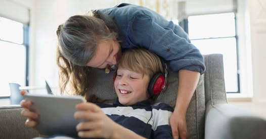 fiatal fiú a fején fejhallgatóval tabpn játszik valamit, egy középkorú nő fölé hajol